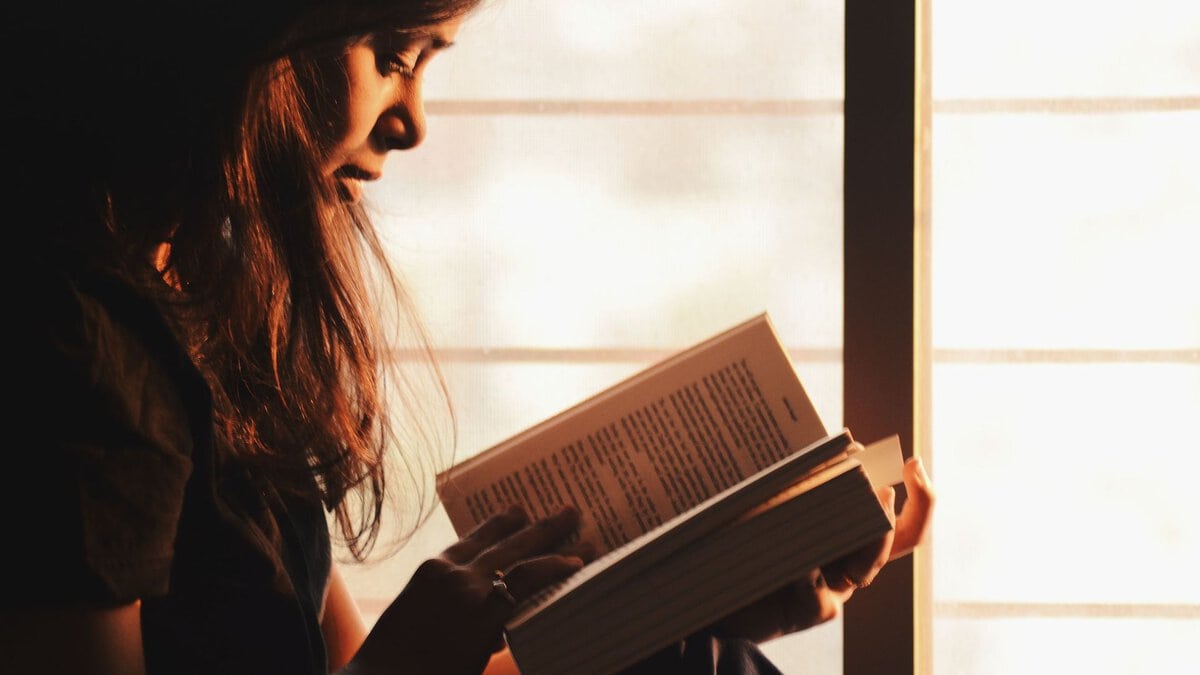 A woman reads a book beside a sunlit window.