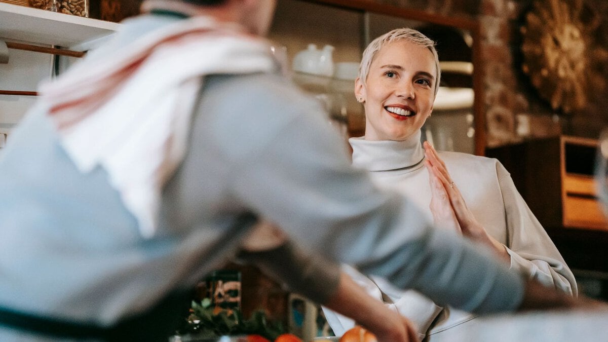 A woman smiles toward a man preparing food at a kitchen counter.