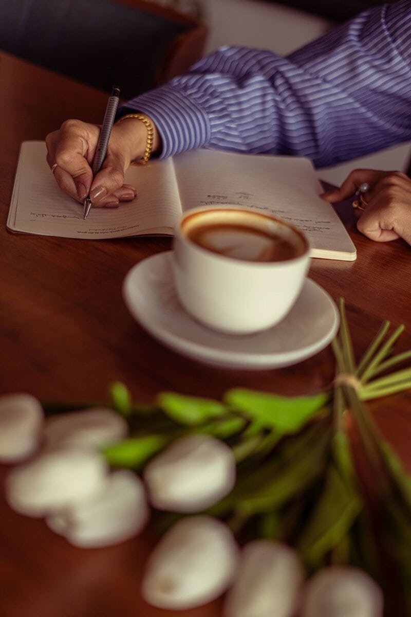 Woman writing in a journal beside coffee and flowers