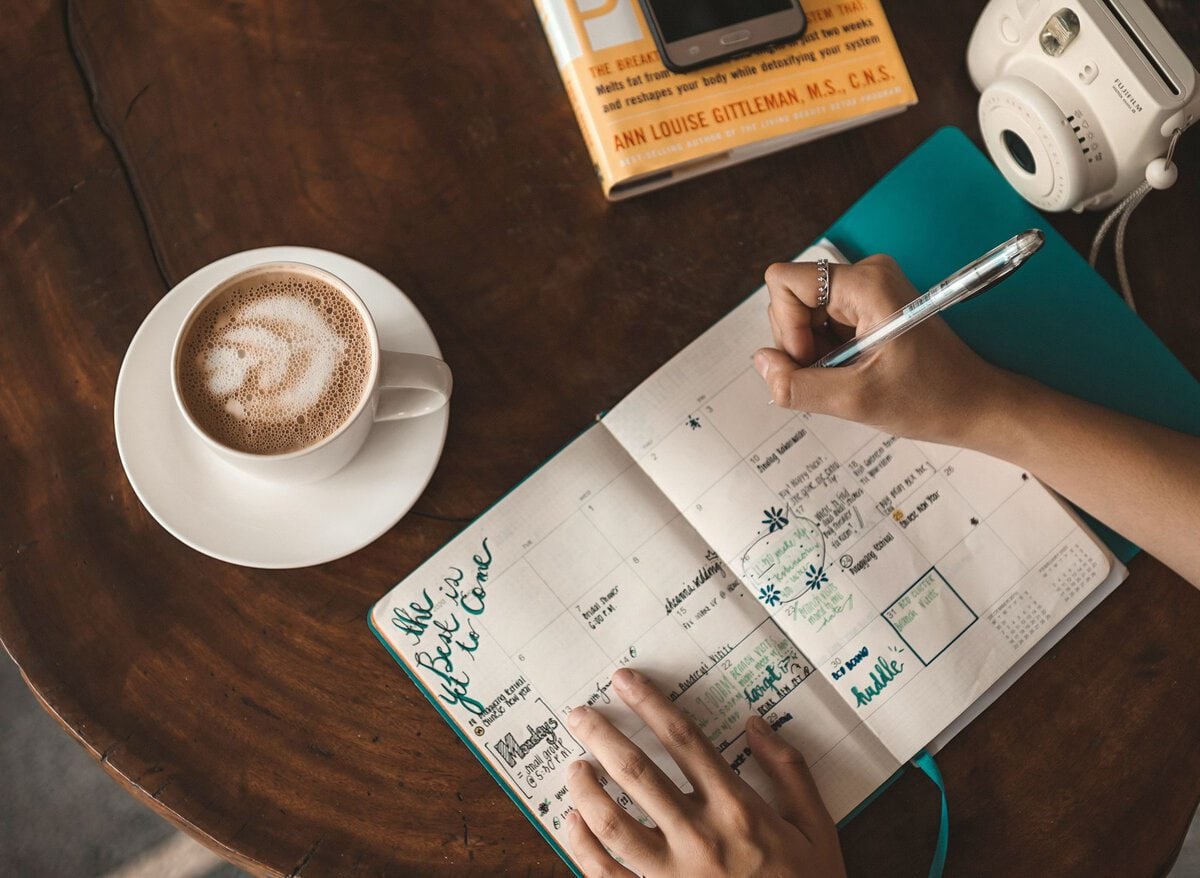 Hands writing plans in a notebook beside coffee